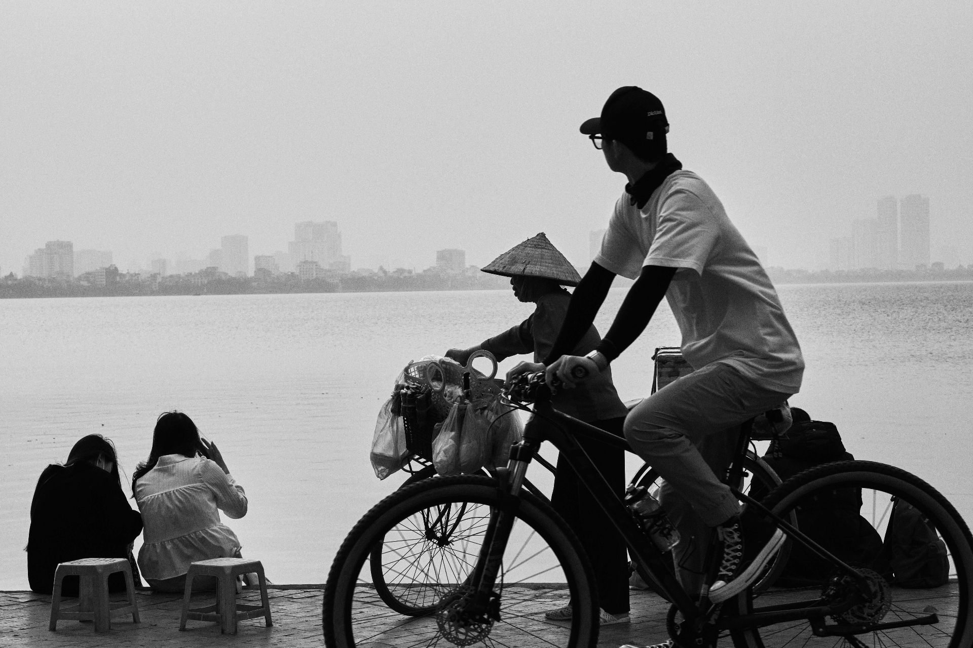 Persone in bicicletta e sedute in riva a un lago in Vietnam, in bianco e nero — incontri autentici e vita quotidiana.